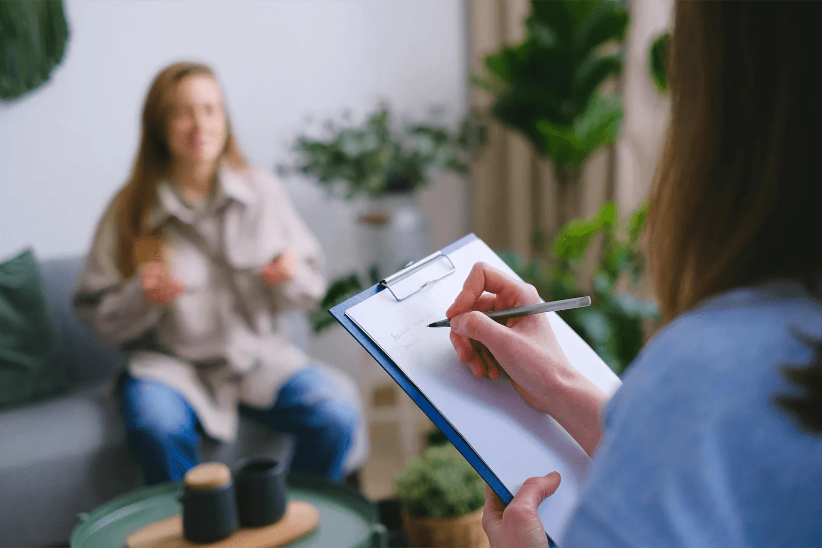 Counseling session captured from therapist's perspective, showing clipboard notes being taken in cozy office setting with greenery