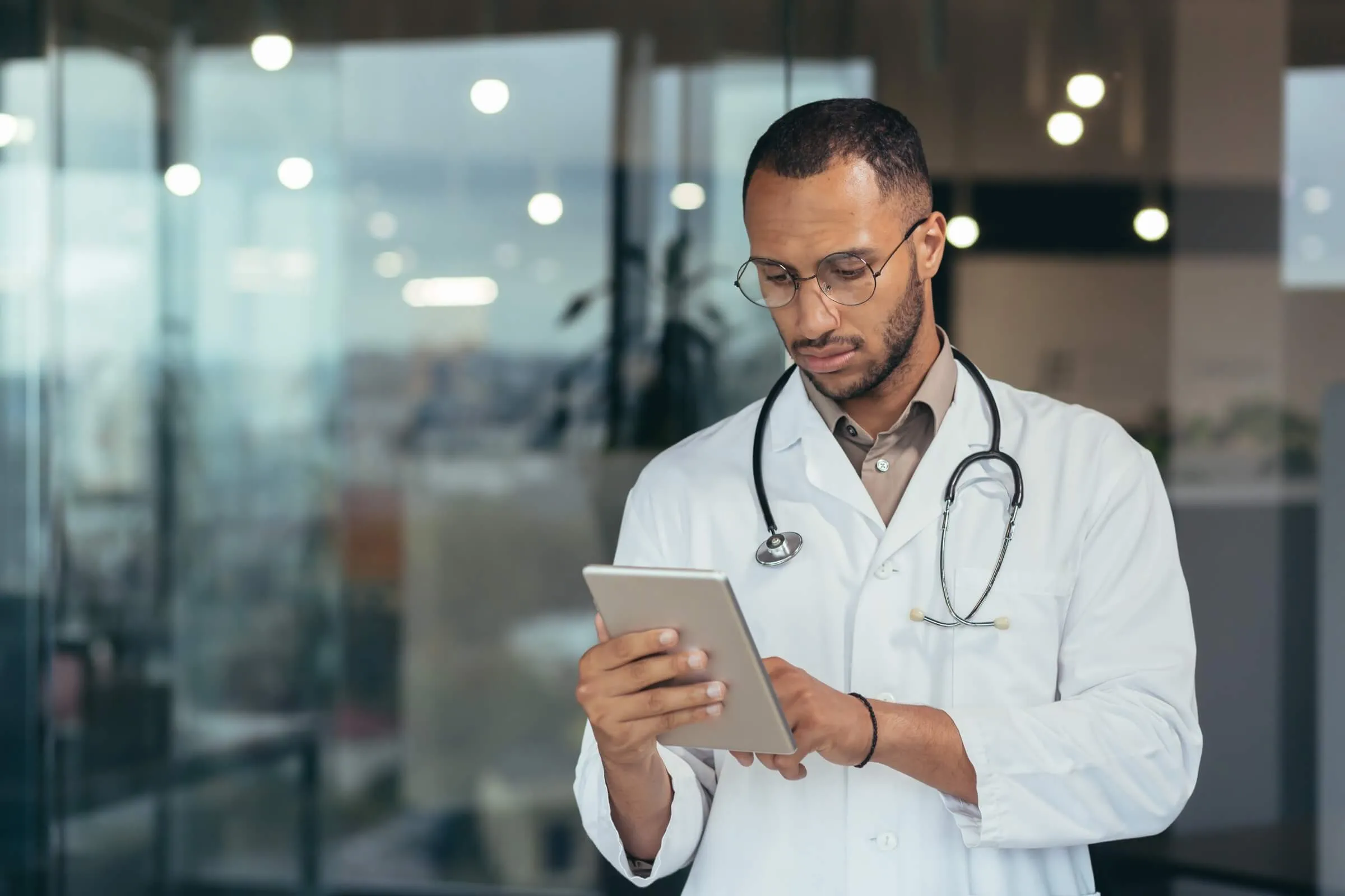 Doctor in white coat with stethoscope checking medical information on tablet in hospital corridor.