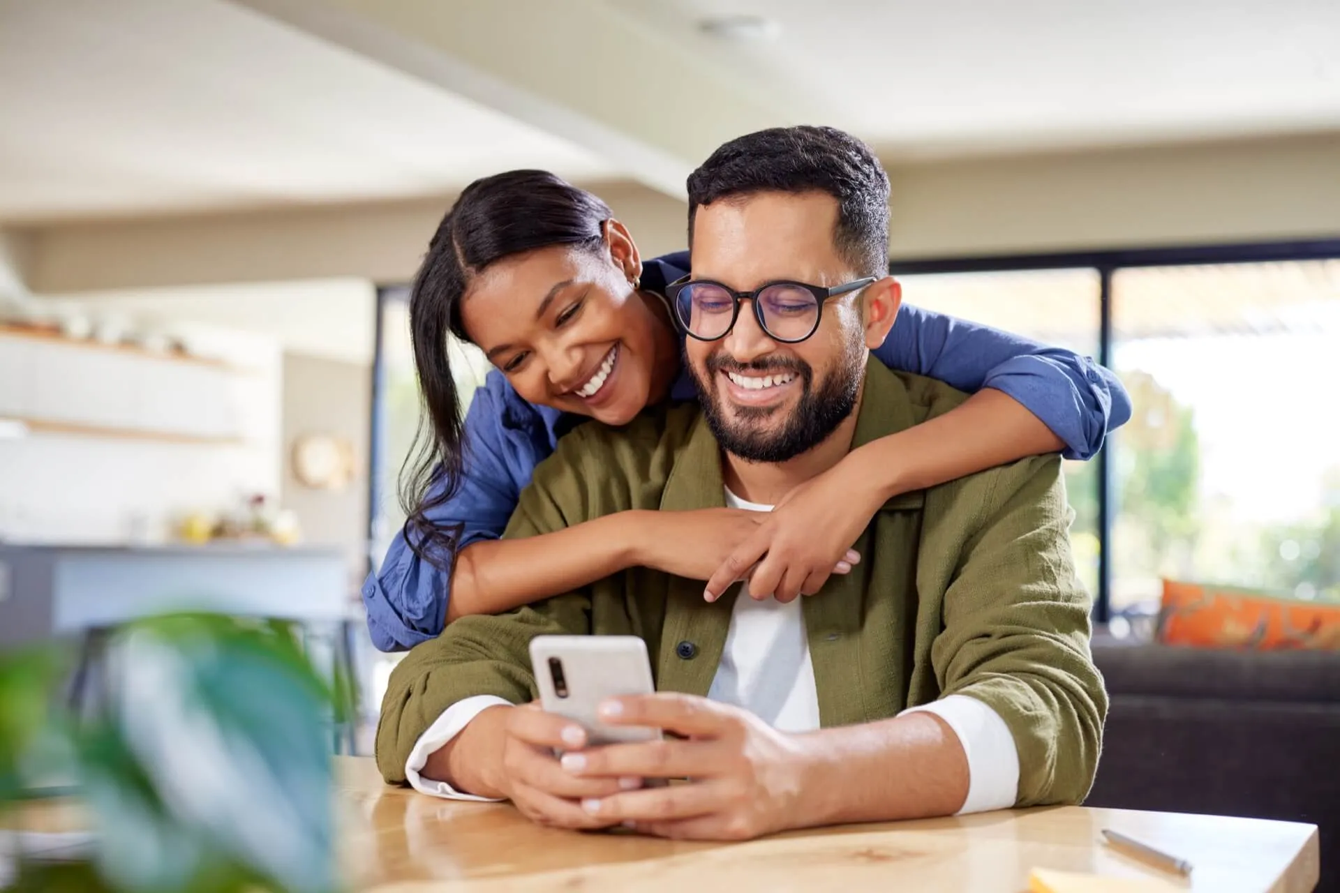 A joyful couple sitting at a table, with the woman playfully hugging the man from behind. The man is holding a smartphone and smiling, while the woman is looking at him with a bright smile. The background features a well-lit room with plants and a cozy atmosphere.