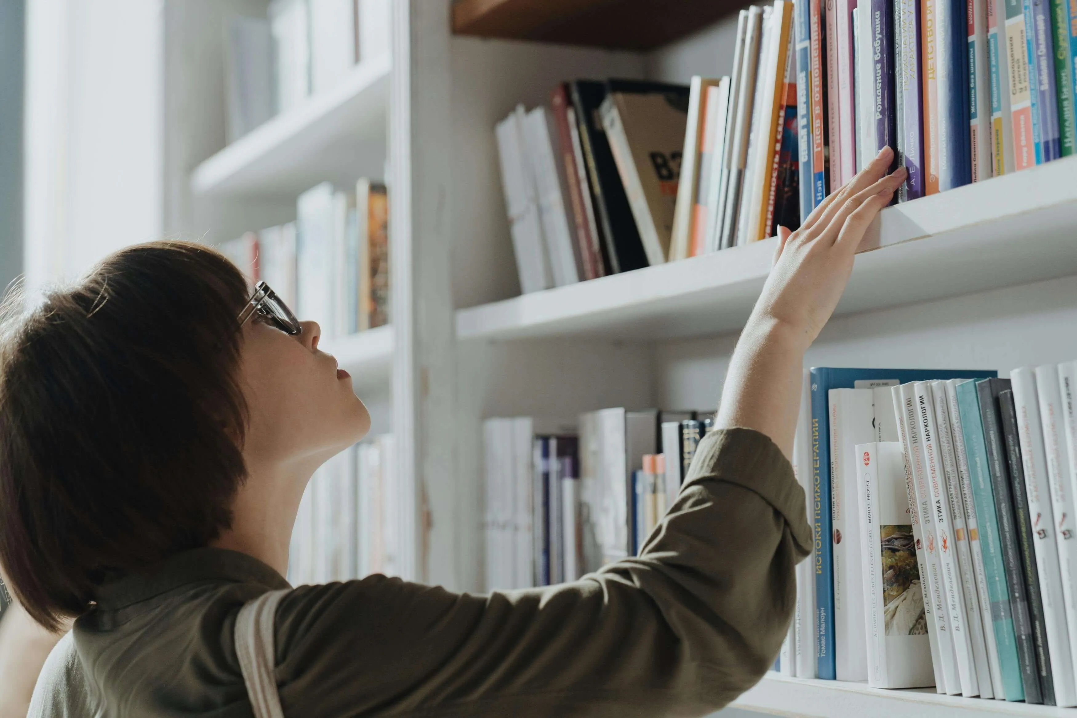 Person reaching for book on white bookshelf, wearing olive green jacket and glasses in soft natural lighting.