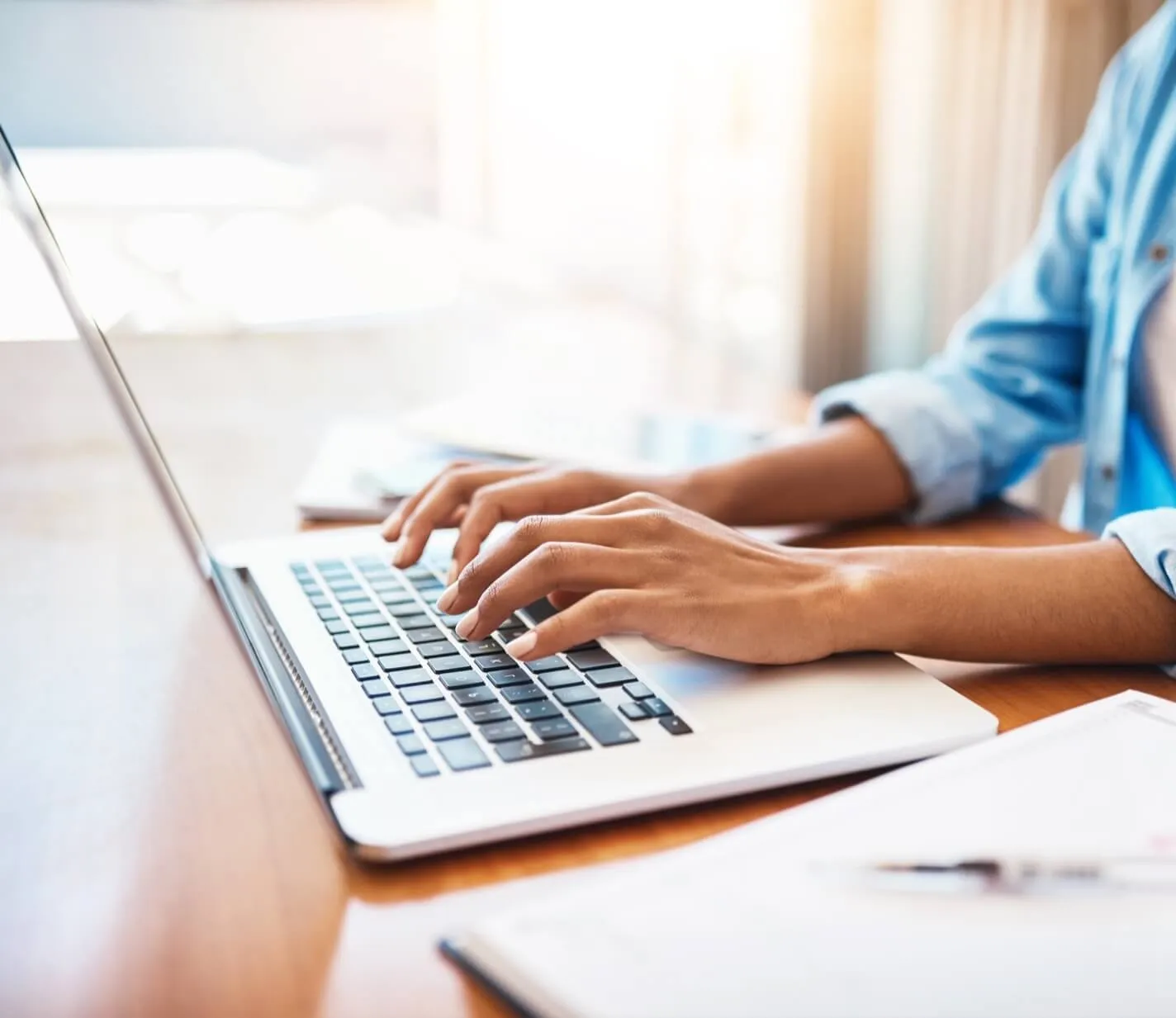 A close-up view of a person's hands typing on a laptop keyboard. The individual is wearing a light blue denim shirt, and the laptop is placed on a wooden table with some papers visible in the background