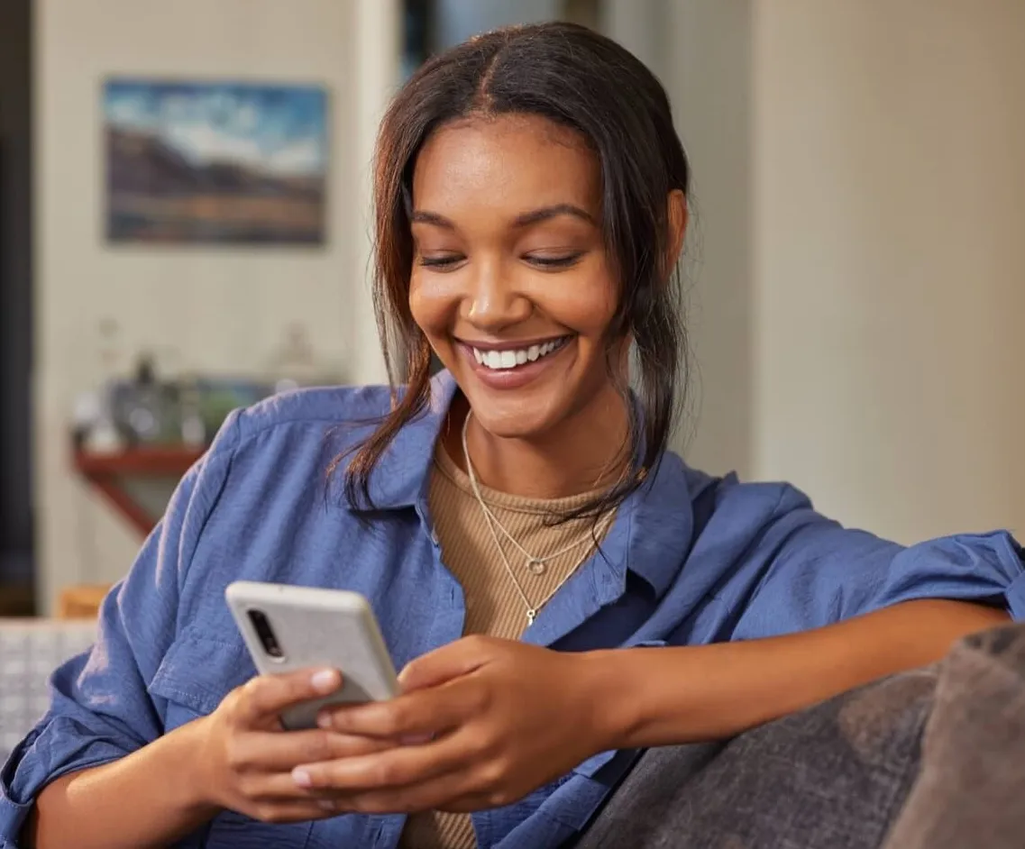 Woman relaxing on couch while using a smartphone, wearing blue denim shirt and beige top, smiling warmly